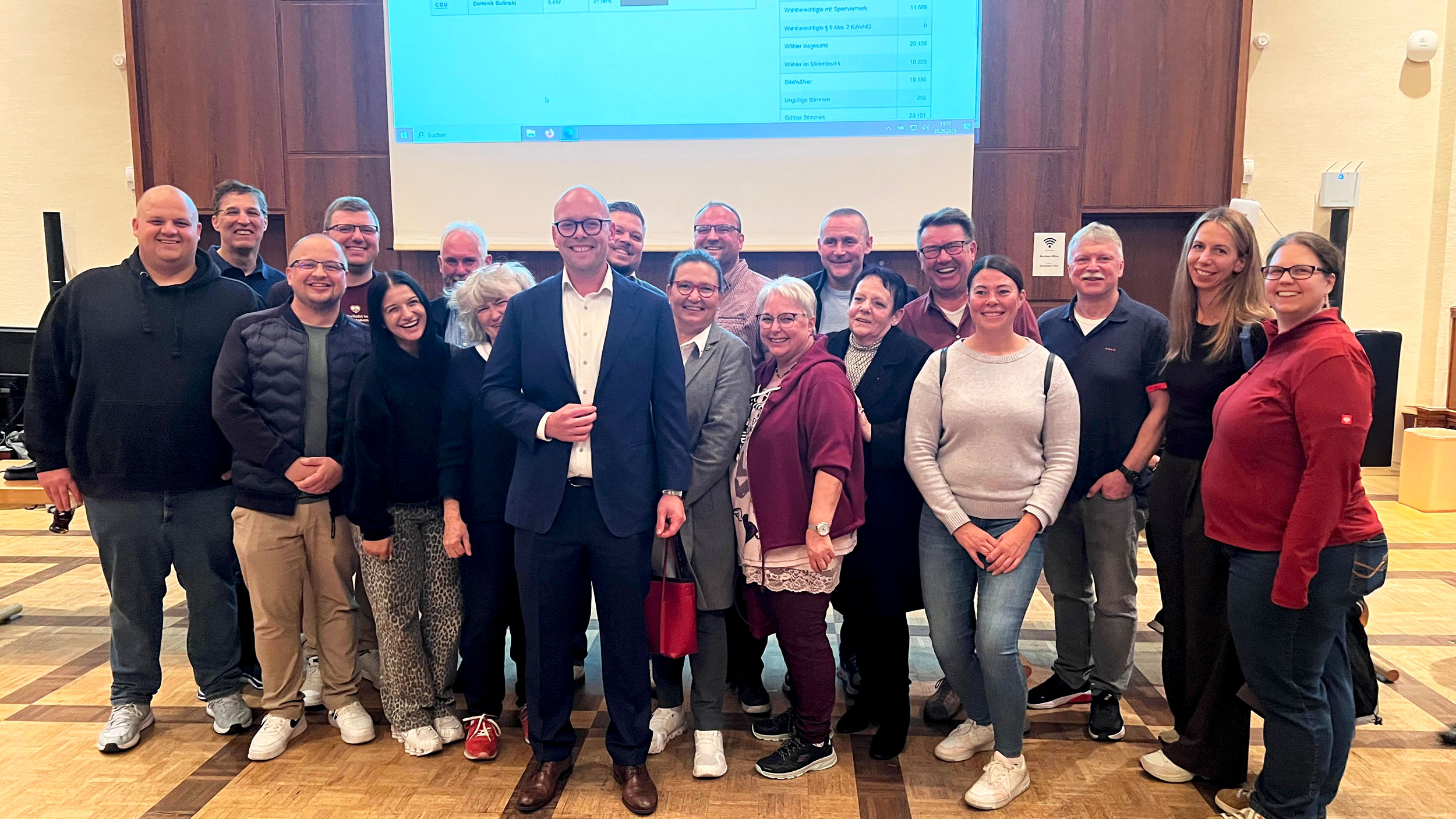 Teile der Fraktionsmitglieder stehen mit dem neugewählten Bürgermeister vor einer Leinwand, auf der das Endergebnis der Bürgermeister-Stichwahl gezeigt wird.