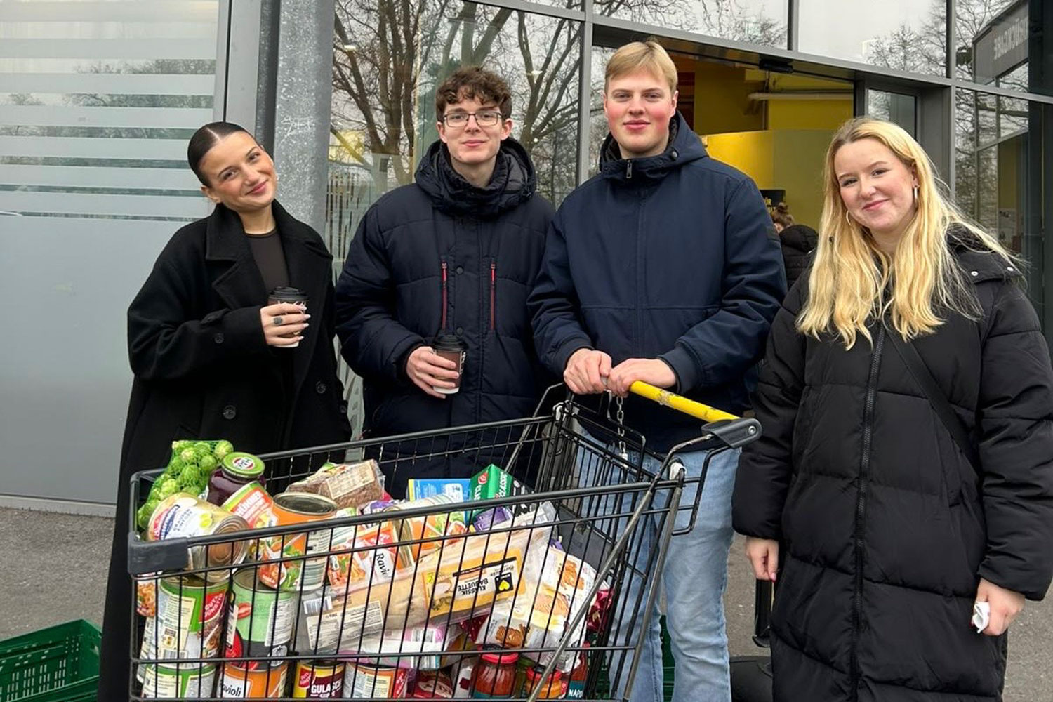 Vier Junge Menschen stehen hinter einem vollbepackten Einkaufswagen.